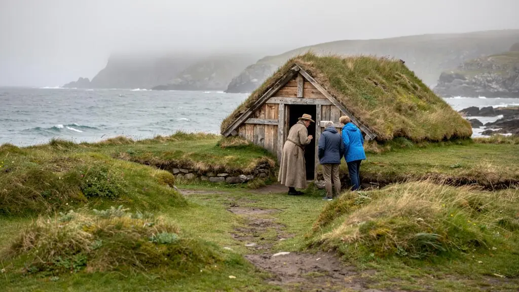 Vue du site archéologique de L'Anse aux Meadows avec des buttes d'herbe marquant les ruines nordiques, une maison en tourbe reconstruite et des visiteurs accompagnés d'un interprète sur le littoral de Terre-Neuve.