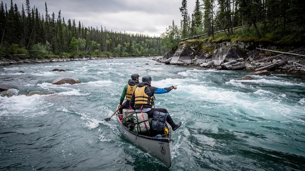 Deux pagayeurs en canot chargé descendent une grande rivière du Nord canadien aux eaux hautes, entourée de forêt d'épinettes et de berges rocheuses.