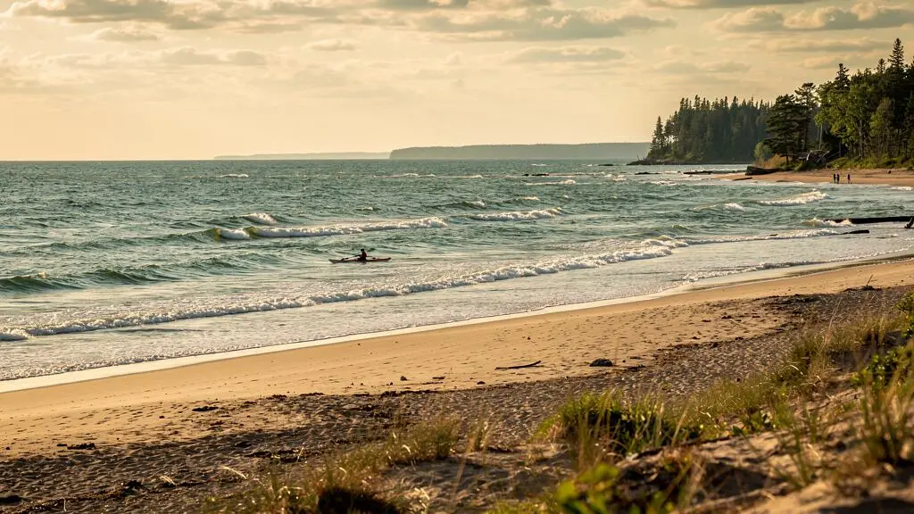 Plage immense au bord d'un grand lac canadien avec des vagues à crêtes blanches et un kayakiste pagayant au loin sous une lumière de fin d'après-midi
