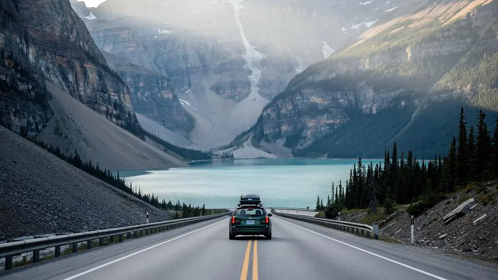 Une voiture circule sur une route panoramique tracée au fond d'une vaste vallée glaciaire en U bordée de parois rocheuses, avec un lac turquoise visible au loin dans les Rocheuses de l'Alberta.